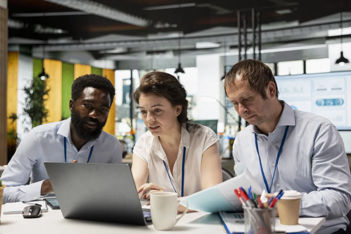 Three colleagues with staff badges reviewing documents on a laptop in an open-plan office